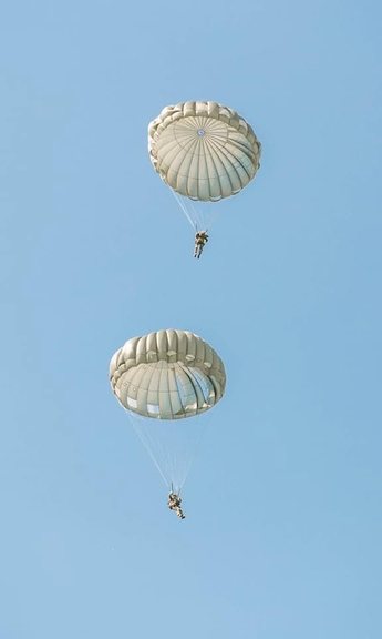 Two paratroopers descending under round parachute canopies against a clear blue sky.