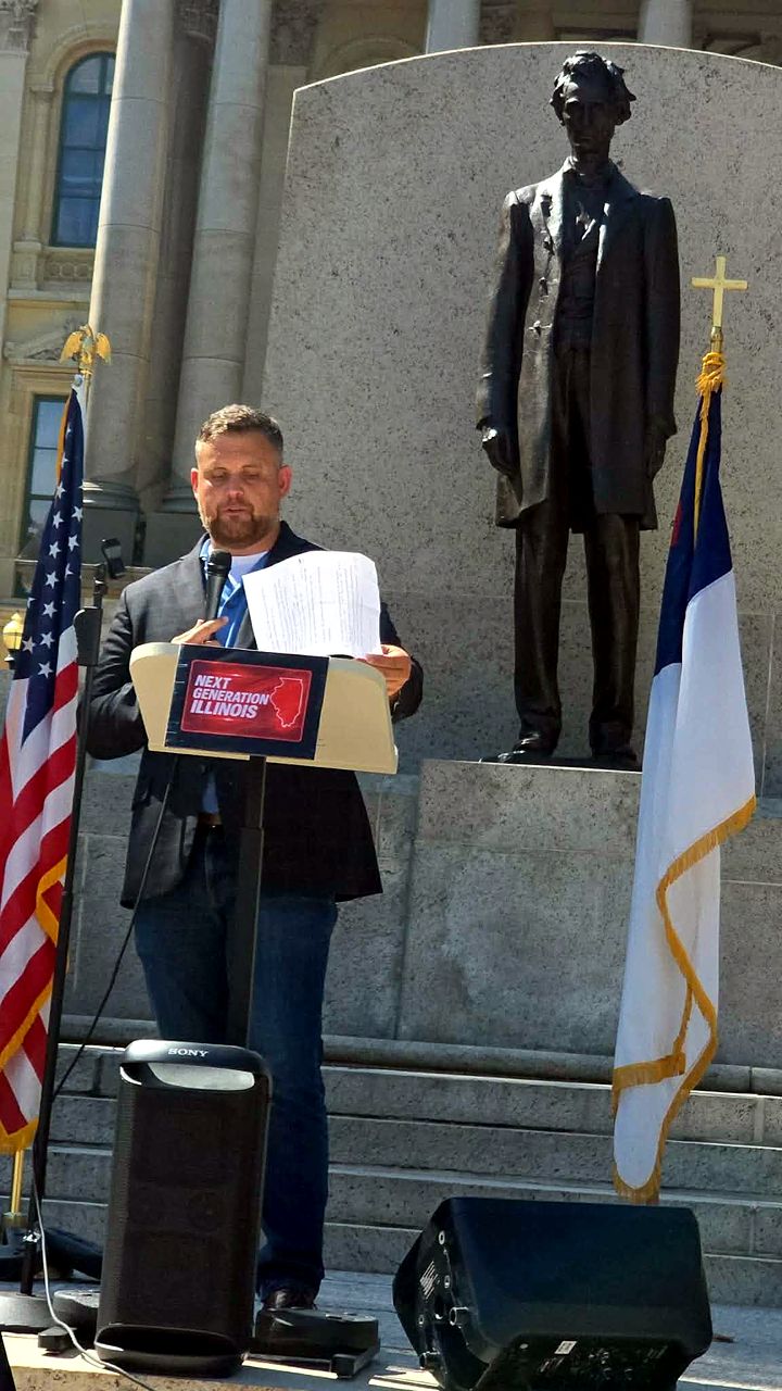 Cody Rose speaking at a podium on the Illinois State Capitol steps, the Lincoln statue and flags behind him.