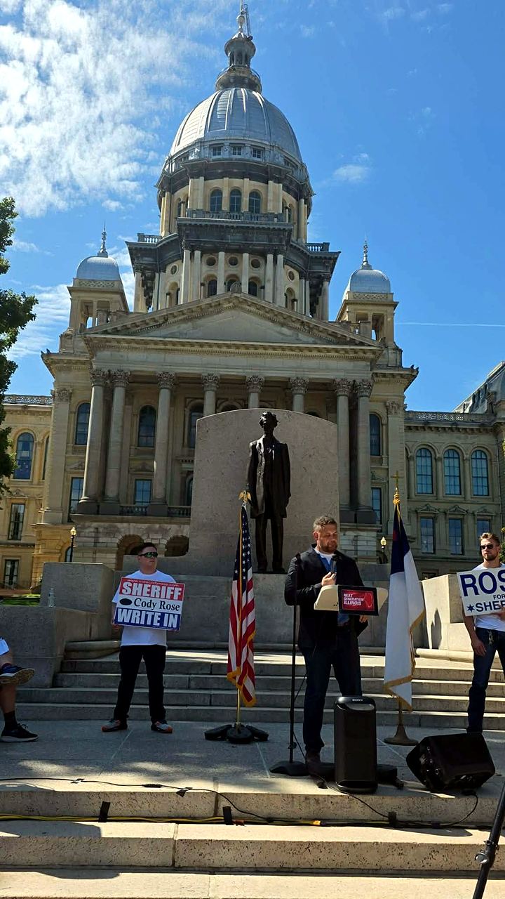 Rally on the Illinois State Capitol steps beside the Lincoln statue, supporters holding Cody Rose write-in signs behind the flags and podium.