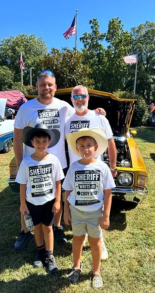 Cody Rose with three boys in Rose for Sheriff write-in t-shirts in front of a yellow car at an outdoor car show.