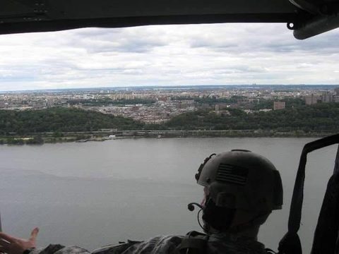 View from the door of a helicopter over the Hudson River, a crew member in a flag-marked flight helmet in the foreground and the New York City skyline beyond.
