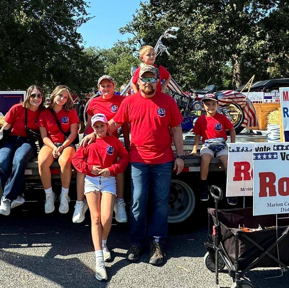 Cody Rose with family and kids in matching red shirts on a parade float, Vote Rose signs stacked alongside.