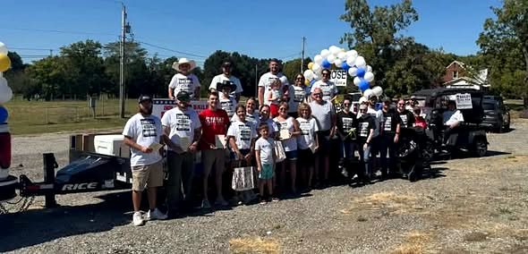 A large group of volunteers in campaign t-shirts standing with signs and balloons beside a trailer before a parade.
